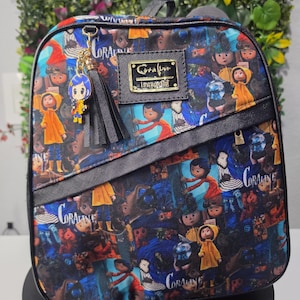 May include: A backpack featuring an all-over print of Coraline characters and scenes in blue, yellow, and orange. It includes a black diagonal front pocket, a black tassel, a gold-toned Coraline charm, and a rectangular metal logo plate reading Coraline.
