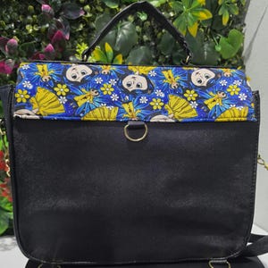 Puede incluir: Un bolso de mano negro con asa superior y cierre de solapa que presenta un panel de tela azul estampado con un patr&oacute;n repetitivo de personajes de dibujos animados con vestidos amarillos, flores blancas y alas azules, con anillas de metal dorado.