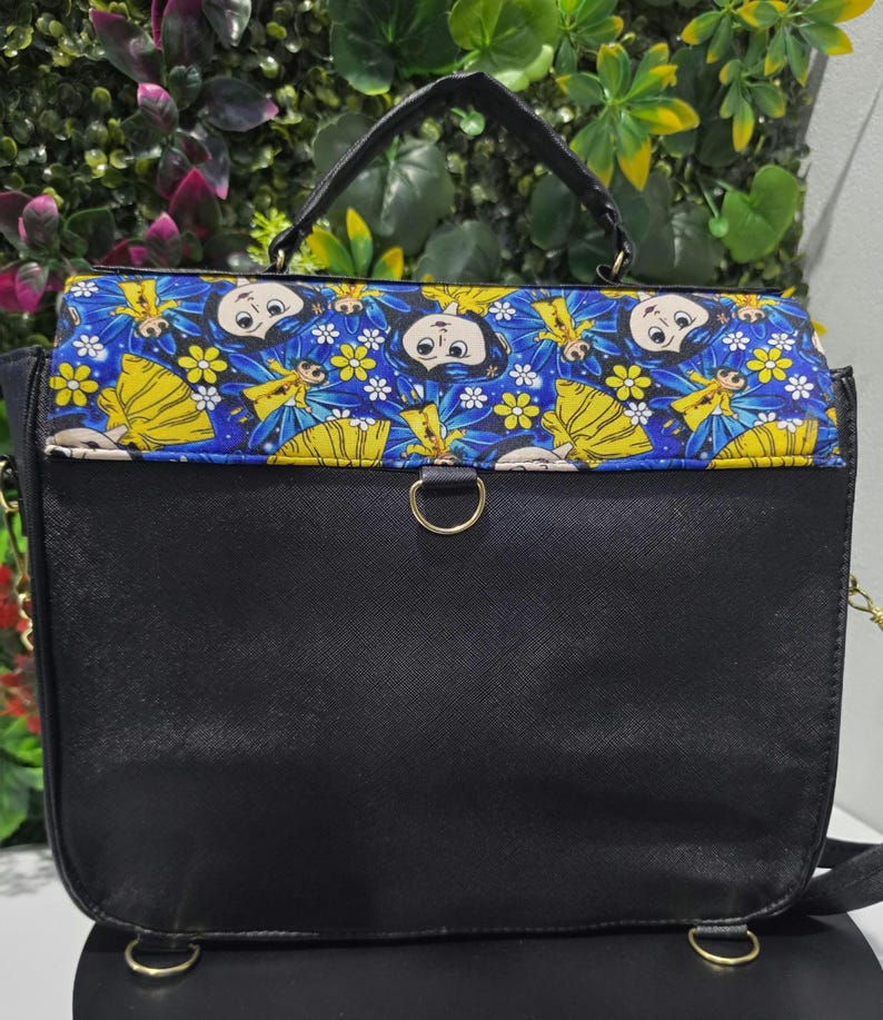Puede incluir: Un bolso de mano negro con asa superior y cierre de solapa que presenta un panel de tela azul estampado con un patr&oacute;n repetitivo de personajes de dibujos animados con vestidos amarillos, flores blancas y alas azules, con anillas de metal dorado.