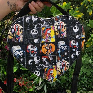 May include: A heart-shaped crossbody bag featuring a black fabric exterior with a repeating comic book style print of characters from The Nightmare Before Christmas, including Jack Skellington and Sally, with black straps and gold-tone hardware.