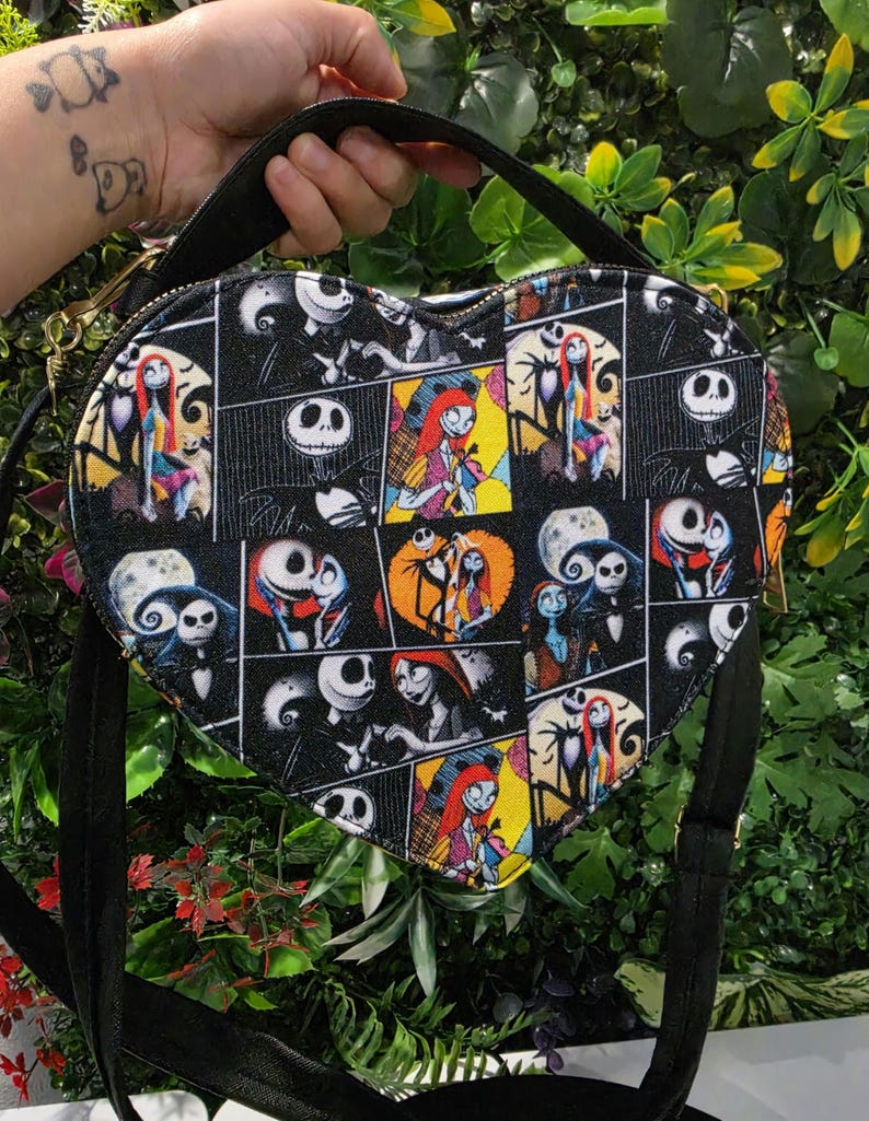 May include: A heart-shaped crossbody bag featuring a black fabric exterior with a repeating comic book style print of characters from The Nightmare Before Christmas, including Jack Skellington and Sally, with black straps and gold-tone hardware.