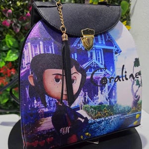May include: A structured backpack featuring a graphic print of the character Coraline standing in front of a house. The bag has a black top flap with a gold-tone metal clasp, a top handle, and a black tassel charm attached to a gold-tone chain.