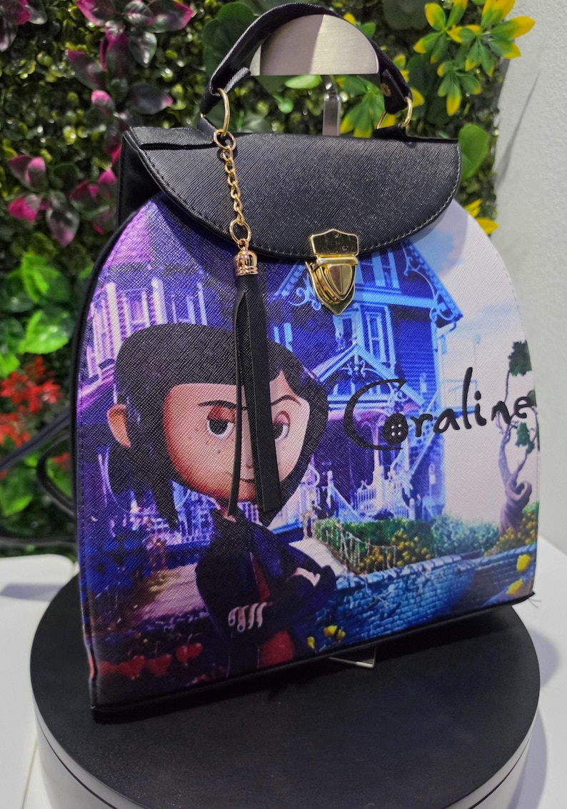 May include: A structured backpack featuring a graphic print of the character Coraline standing in front of a house. The bag has a black top flap with a gold-tone metal clasp, a top handle, and a black tassel charm attached to a gold-tone chain.