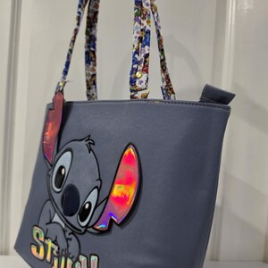 May include: A periwinkle blue tote bag featuring a large graphic of the character Stitch with iridescent accents on the ears and the name Stitch printed in holographic letters. The bag has two patterned shoulder straps with a colorful character print.