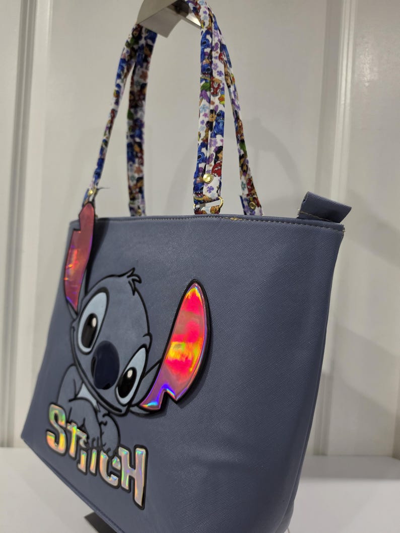 May include: A periwinkle blue tote bag featuring a large graphic of the character Stitch with iridescent accents on the ears and the name Stitch printed in holographic letters. The bag has two patterned shoulder straps with a colorful character print.