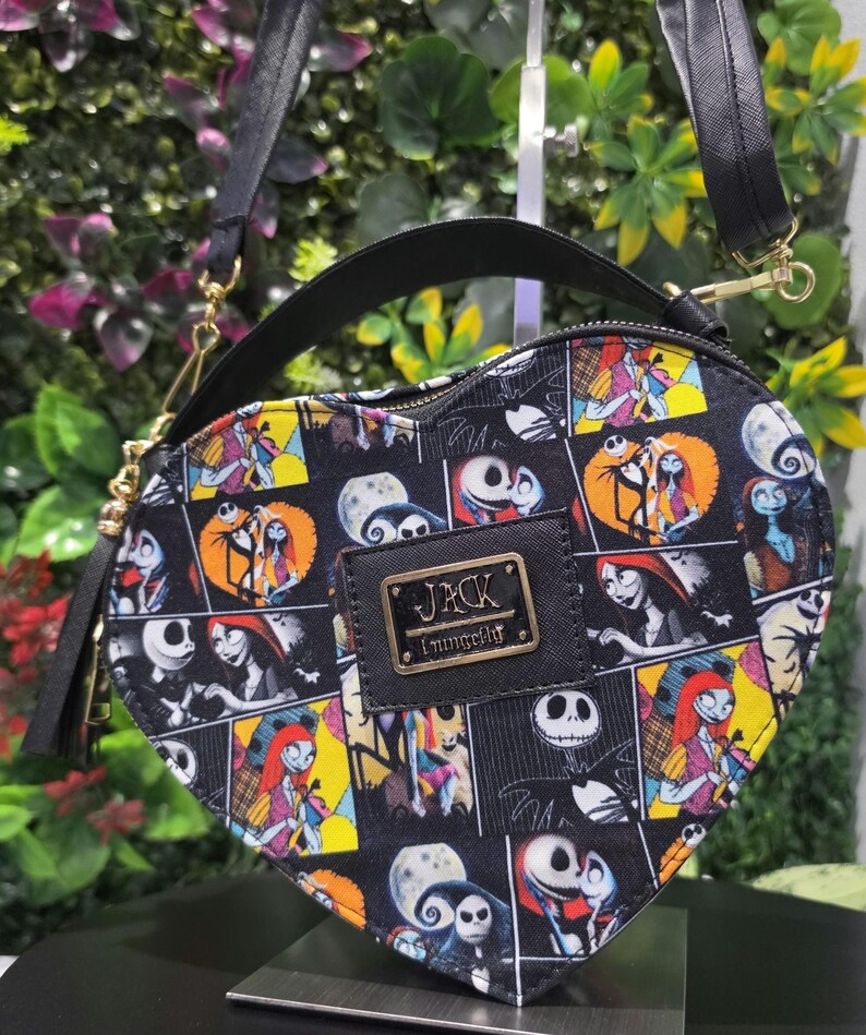 May include: A heart-shaped crossbody bag featuring a black background with a colorful comic-style print of Jack Skellington and Sally from The Nightmare Before Christmas. The bag includes a black top handle, a shoulder strap, and a gold metal plaque with the text JACK Loungefly.