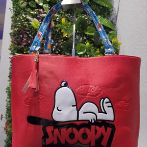 May include: A red tote bag featuring a white Snoopy character graphic with the text SNOOPY in black letters. The bag includes blue patterned handles with small character prints and a red tassel charm attached to one side of the handle hardware.