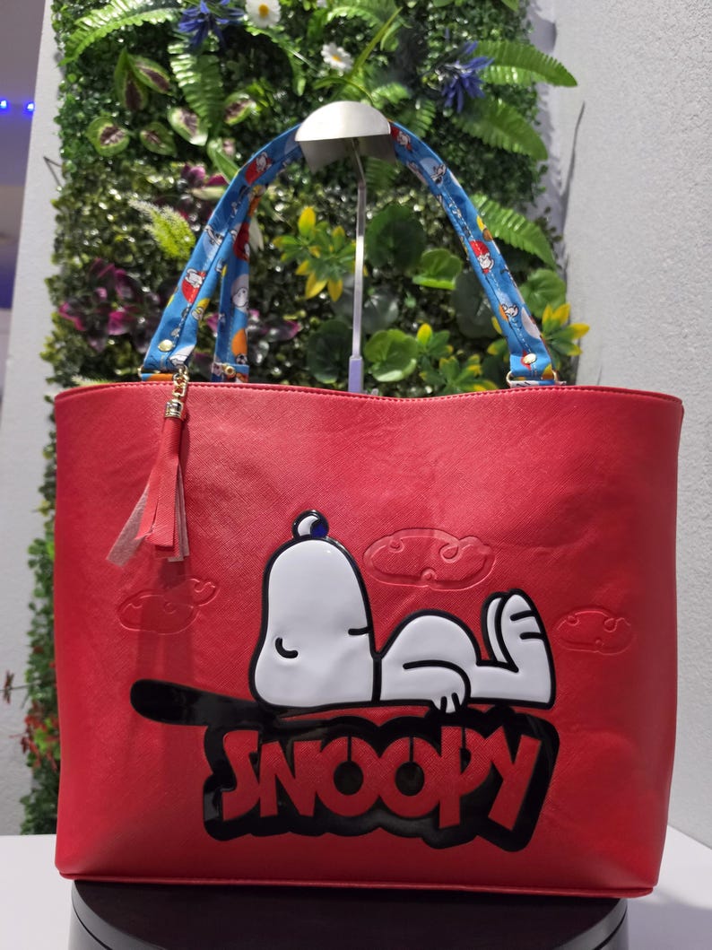 May include: A red tote bag featuring a white Snoopy character graphic with the text SNOOPY in black letters. The bag includes blue patterned handles with small character prints and a red tassel charm attached to one side of the handle hardware.