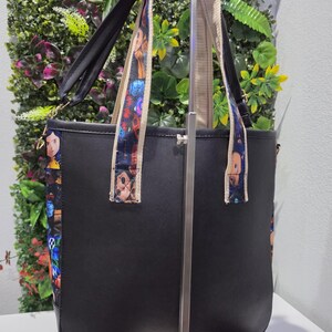 May include: A black tote bag featuring side panels and shoulder straps decorated with a colorful character print. The bag has a structured design with a solid black front and back, and includes a long adjustable strap attached to the sides.