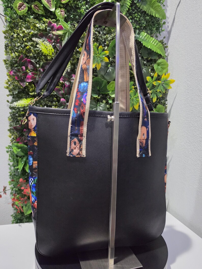 May include: A black tote bag featuring side panels and shoulder straps decorated with a colorful character print. The bag has a structured design with a solid black front and back, and includes a long adjustable strap attached to the sides.