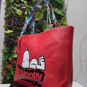 May include: A bright red tote bag featuring a large graphic of the character Snoopy lying down with the text SNOOPY printed in bold black letters below. The bag includes two blue patterned shoulder straps and a small red tassel attached to the side.