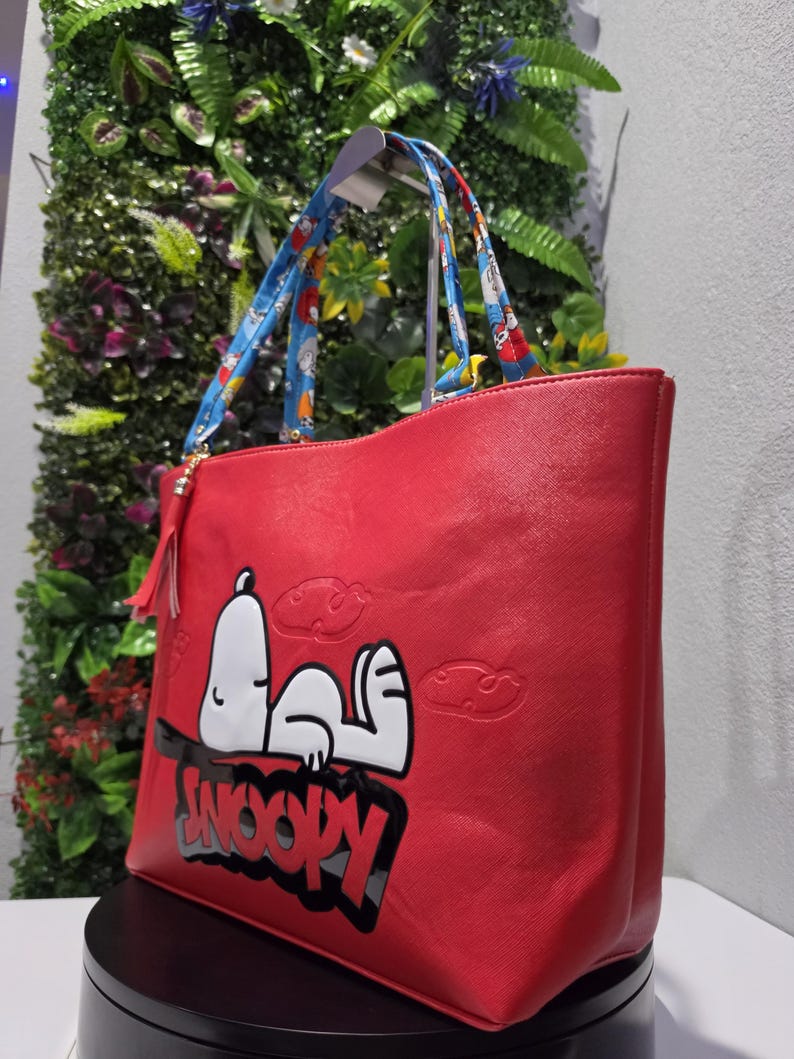 May include: A bright red tote bag featuring a large graphic of the character Snoopy lying down with the text SNOOPY printed in bold black letters below. The bag includes two blue patterned shoulder straps and a small red tassel attached to the side.