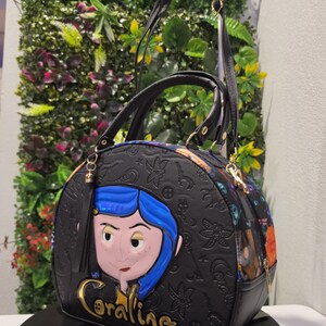 May include: A black dome-shaped handbag featuring a raised, colorful character applique of Coraline with blue hair. The bag has an embossed pattern of keys, stars, and buttons, with the name Coraline in gold lettering and a black tassel attachment.