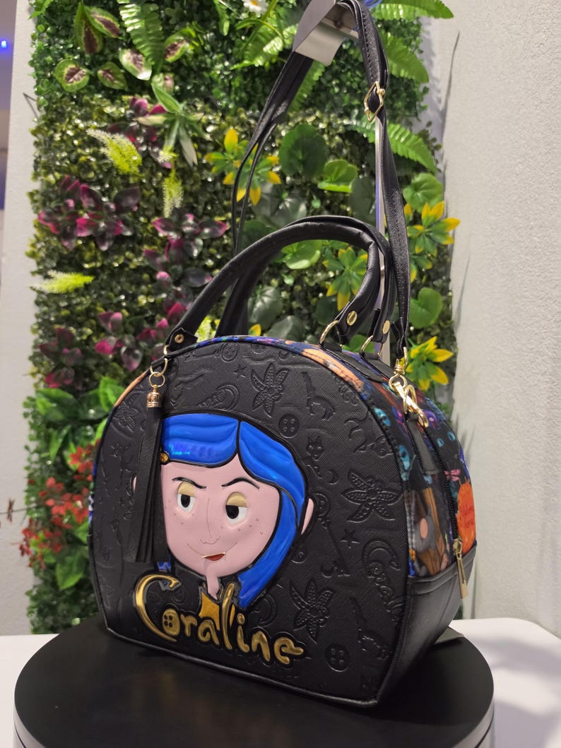 May include: A black dome-shaped handbag featuring a raised, colorful character applique of Coraline with blue hair. The bag has an embossed pattern of keys, stars, and buttons, with the name Coraline in gold lettering and a black tassel attachment.