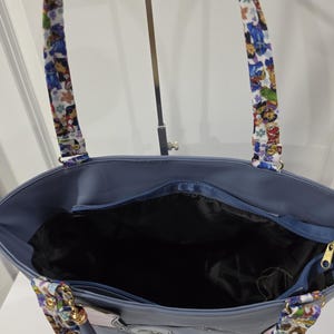 May include: A blue tote bag with a black interior lining and a zippered top closure. The bag features two shoulder straps decorated with a colorful cartoon character print. The exterior is a solid blue color with gold-toned metal hardware.