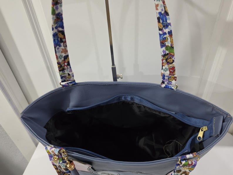 May include: A blue tote bag with a black interior lining and a zippered top closure. The bag features two shoulder straps decorated with a colorful cartoon character print. The exterior is a solid blue color with gold-toned metal hardware.