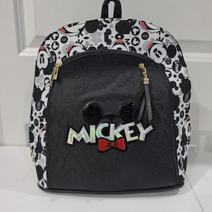 May include: A black and white backpack featuring a repeating Mickey Mouse pattern on the sides and top. The front pocket is solid black with a debossed Mickey icon pattern, a central Mickey Mouse head emblem, a red bow, and the word MICKEY in silver.