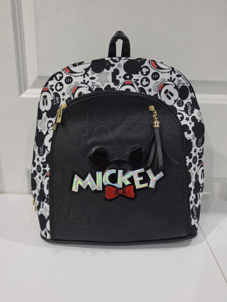 May include: A black and white backpack featuring a repeating Mickey Mouse pattern on the sides and top. The front pocket is solid black with a debossed Mickey icon pattern, a central Mickey Mouse head emblem, a red bow, and the word MICKEY in silver.