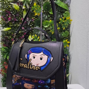 以下が含まれることがあります： フロントフラップに青い髪のコララインのキャラクターアップリケと金色の文字でCoralineと書かれた黒いハンドバッグ。下部にはコララインのキャラクターと黒猫が描かれたカラフルなプリントがあり、タッセルチャームが付いています。