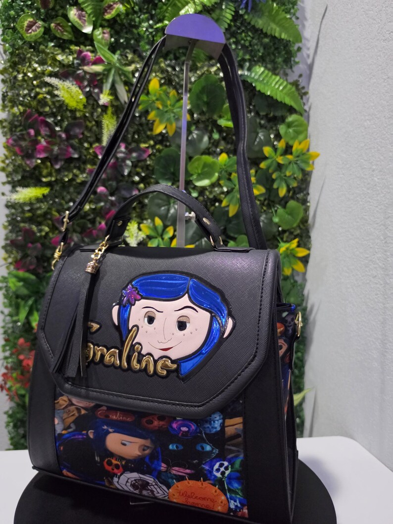 以下が含まれることがあります： フロントフラップに青い髪のコララインのキャラクターアップリケと金色の文字でCoralineと書かれた黒いハンドバッグ。下部にはコララインのキャラクターと黒猫が描かれたカラフルなプリントがあり、タッセルチャームが付いています。