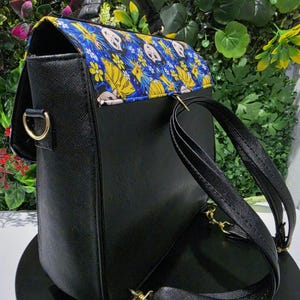 Puede incluir: Una mochila negra con acabado texturizado y asa superior. La solapa presenta una tela azul vibrante con un patr&oacute;n repetitivo de flores amarillas y rostros de personajes de dibujos animados. Incluye herrajes met&aacute;licos dorados y correas.