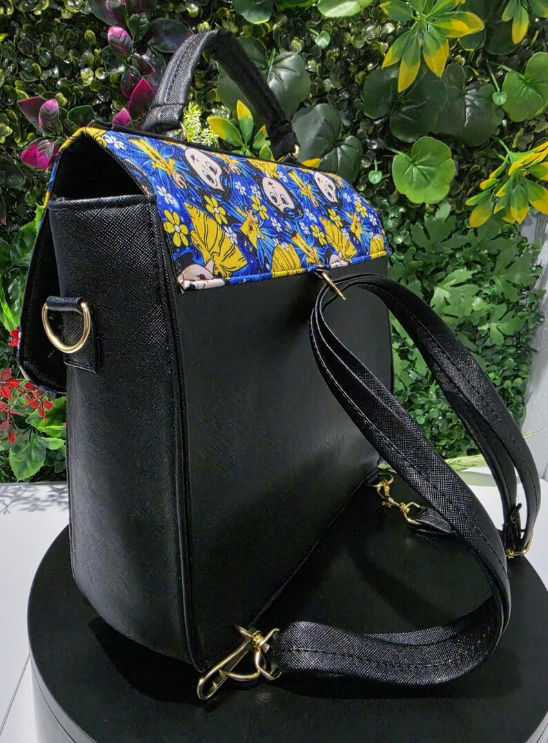 Puede incluir: Una mochila negra con acabado texturizado y asa superior. La solapa presenta una tela azul vibrante con un patr&oacute;n repetitivo de flores amarillas y rostros de personajes de dibujos animados. Incluye herrajes met&aacute;licos dorados y correas.