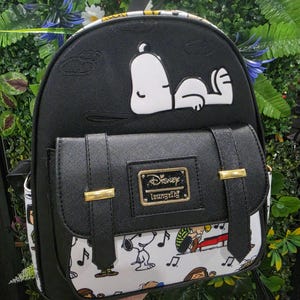 Puede incluir: Mochila negra con Snoopy de Peanuts. La mochila tiene un bolsillo con solapa con detalles dorados y un diseño en blanco y negro. El logotipo de Disney Loungefly es visible en la parte delantera.