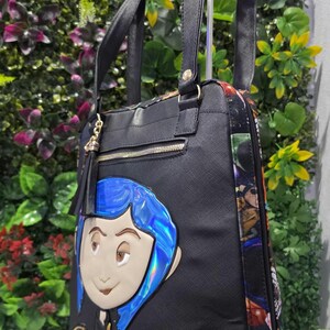 Puede incluir: Un bolso tote negro con una aplicaci&oacute;n del personaje Coraline con cabello azul iridiscente y el nombre Coraline en letras doradas. El bolso tiene correas negras, un bolsillo frontal con cremallera y borla, y paneles laterales con estampado.