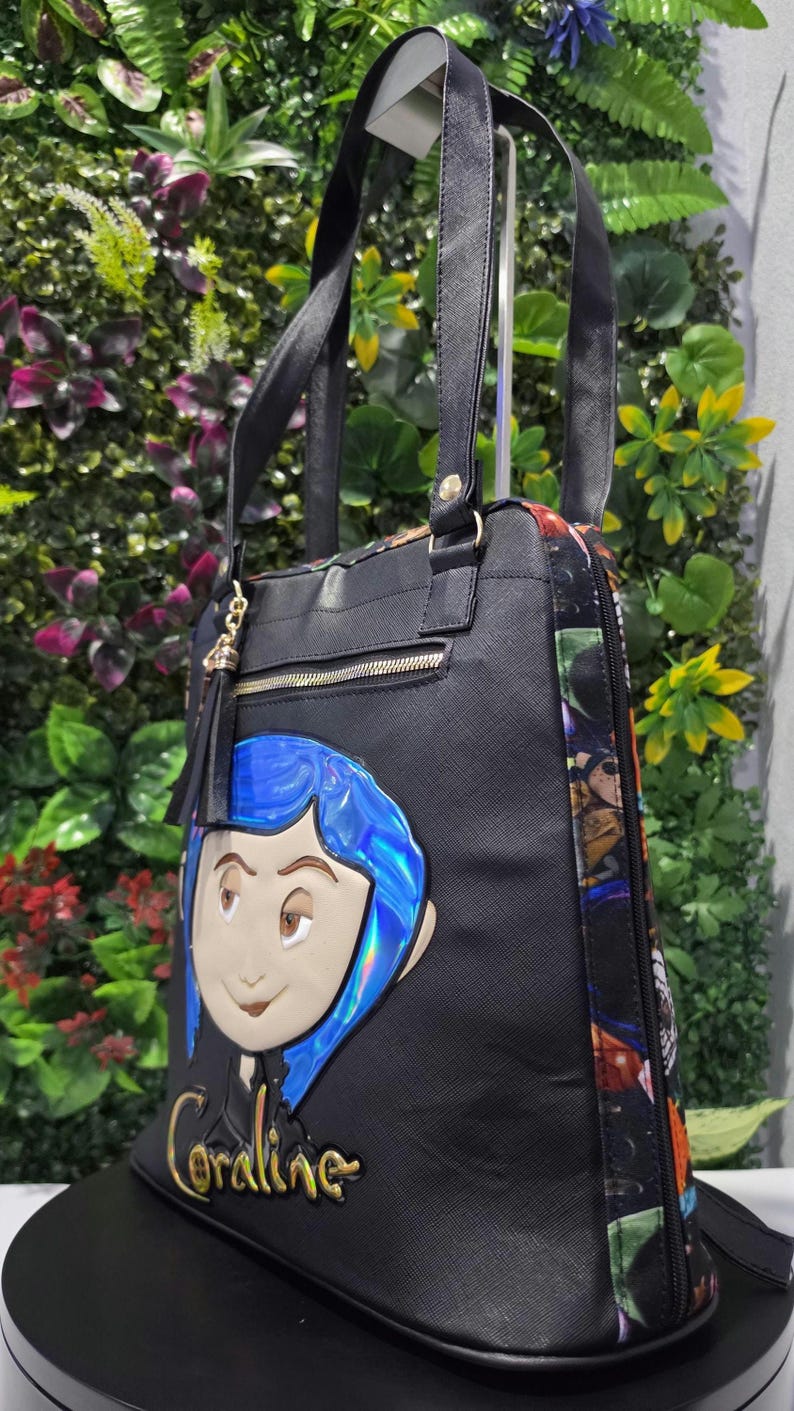 Puede incluir: Un bolso tote negro con una aplicaci&oacute;n del personaje Coraline con cabello azul iridiscente y el nombre Coraline en letras doradas. El bolso tiene correas negras, un bolsillo frontal con cremallera y borla, y paneles laterales con estampado.