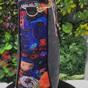 May include: A black tote bag featuring a side panel with a colorful graphic print of characters, spiders, and buttons, including the text Some doors should never be opened. The bag has black trim, gold-tone metal hardware, and dual shoulder straps.