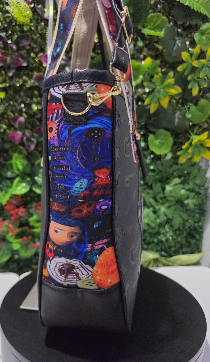 May include: A black tote bag featuring a side panel with a colorful graphic print of characters, spiders, and buttons, including the text Some doors should never be opened. The bag has black trim, gold-tone metal hardware, and dual shoulder straps.