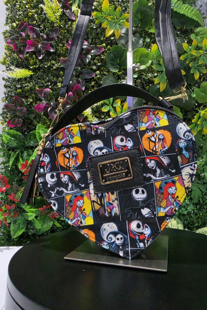May include: A heart-shaped crossbody bag featuring a black background with a colorful collage print of characters from The Nightmare Before Christmas. The bag includes a black top handle, an adjustable shoulder strap, and a gold-toned Jack Loungefly logo.
