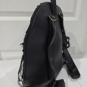 May include: A black backpack with a side profile view showing a smooth black back panel, adjustable black shoulder straps, a top carry handle, and a side zipper closure. The front section features a white and black patterned design.