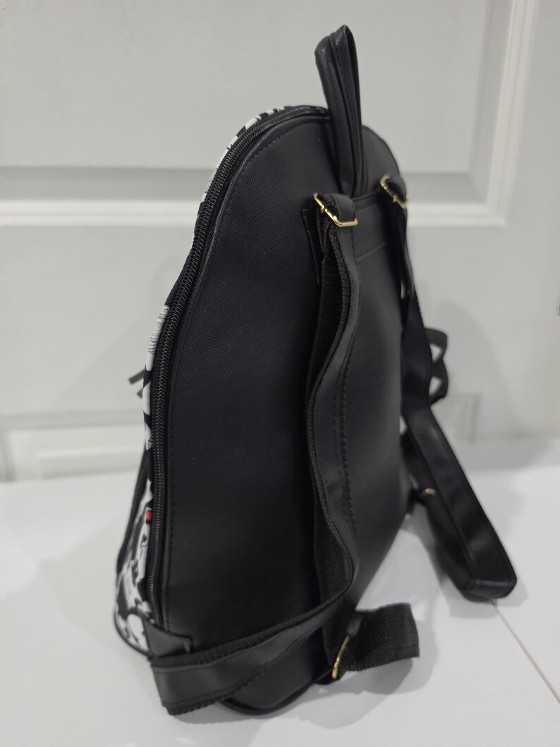 May include: A black backpack with a side profile view showing a smooth black back panel, adjustable black shoulder straps, a top carry handle, and a side zipper closure. The front section features a white and black patterned design.