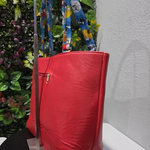 May include: A bright red tote bag with a textured surface and a gold-toned zip on the side. The bag features two shoulder straps made of blue fabric decorated with a repeating cartoon character pattern in yellow, white, and black colours.