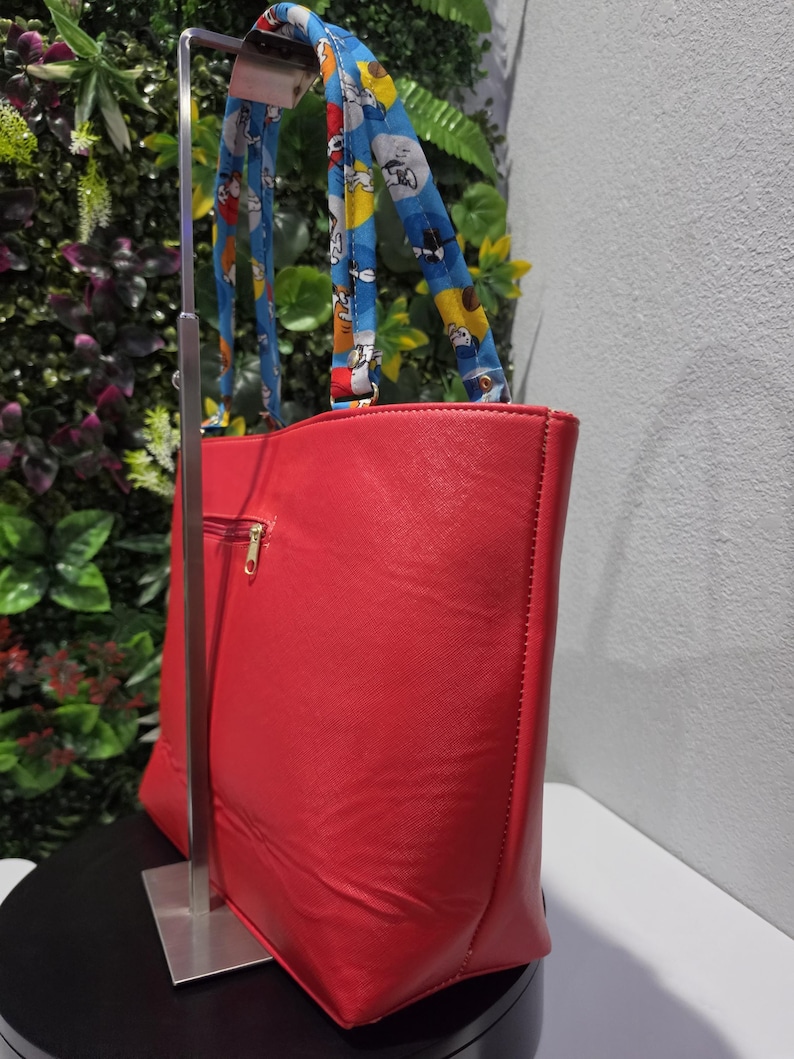 May include: A bright red tote bag with a textured surface and a gold-toned zip on the side. The bag features two shoulder straps made of blue fabric decorated with a repeating cartoon character pattern in yellow, white, and black colours.