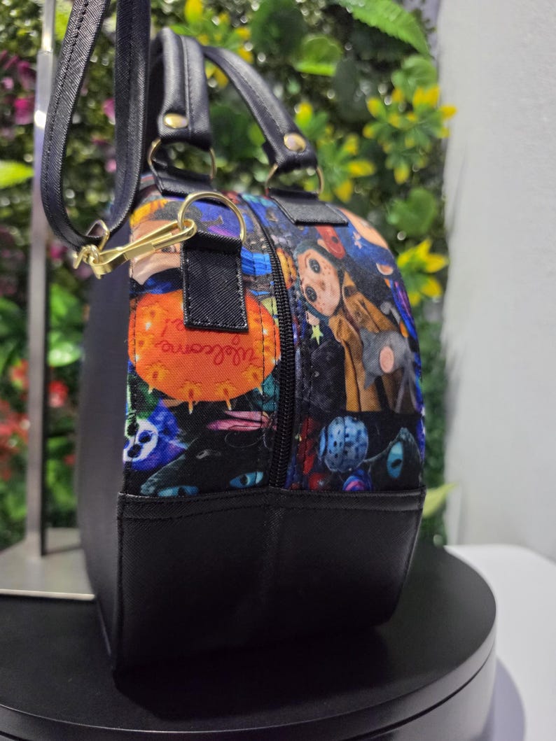 May include: A round black handbag featuring a colorful patterned side panel with illustrations of a character with button eyes, a cat, and an orange sign with the text "Welcome!". The bag has black handles, a zipper closure, and a detachable shoulder strap.