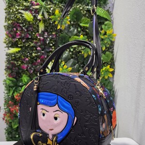 May include: A black dome-shaped handbag featuring a raised Coraline character applique with blue hair, gold lettering reading Coraline, and an embossed pattern of keys and stars. The bag includes black top handles and an adjustable shoulder strap.