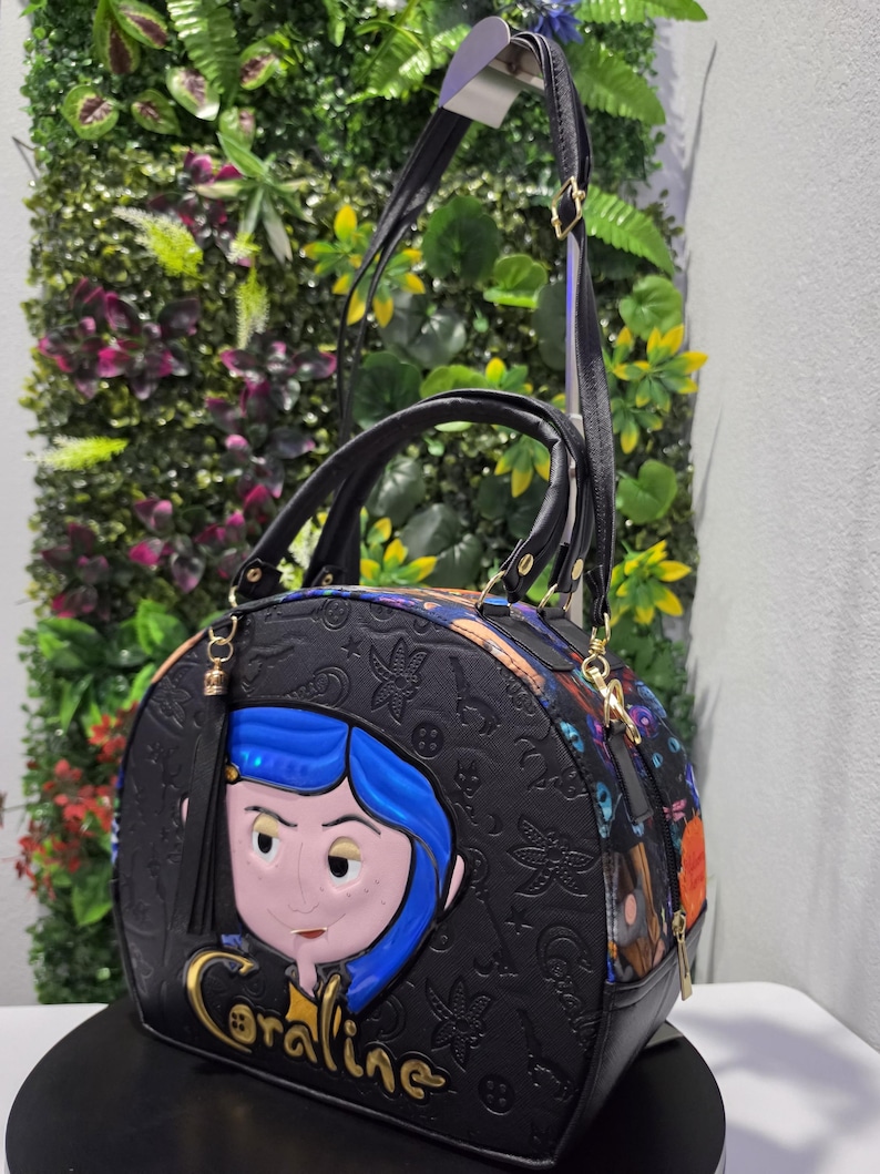 May include: A black dome-shaped handbag featuring a raised Coraline character applique with blue hair, gold lettering reading Coraline, and an embossed pattern of keys and stars. The bag includes black top handles and an adjustable shoulder strap.
