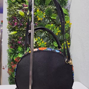 May include: A semi-circular black handbag featuring a colorful patterned trim with skulls and floral motifs. The bag includes a top handle, an adjustable shoulder strap, and gold-toned hardware. The main body is made of a textured black material.