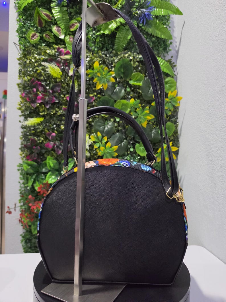 May include: A semi-circular black handbag featuring a colorful patterned trim with skulls and floral motifs. The bag includes a top handle, an adjustable shoulder strap, and gold-toned hardware. The main body is made of a textured black material.