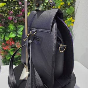 May include: A black backpack with a textured finish, featuring a top handle, adjustable shoulder straps, and gold-toned metal hardware. The bag has a flap closure, a zippered main compartment, and side D-rings for attaching additional straps.