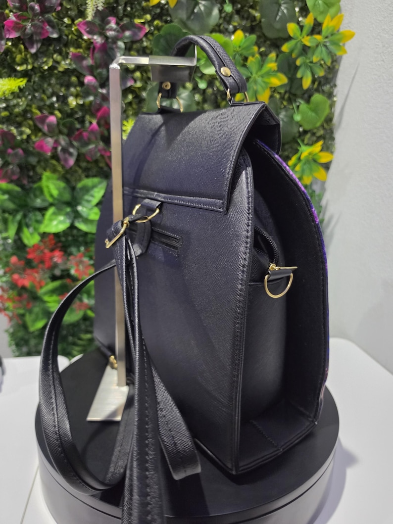 May include: A black backpack with a textured finish, featuring a top handle, adjustable shoulder straps, and gold-toned metal hardware. The bag has a flap closure, a zippered main compartment, and side D-rings for attaching additional straps.