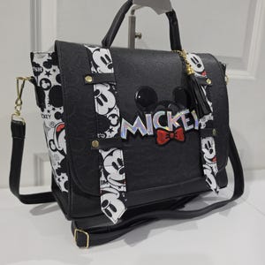 May include: A black satchel bag featuring a debossed pattern, a top handle, and an adjustable shoulder strap. The front flap displays the word MICKEY in iridescent lettering with a red bow, accented by two vertical white strips with black Mickey Mouse graphics.