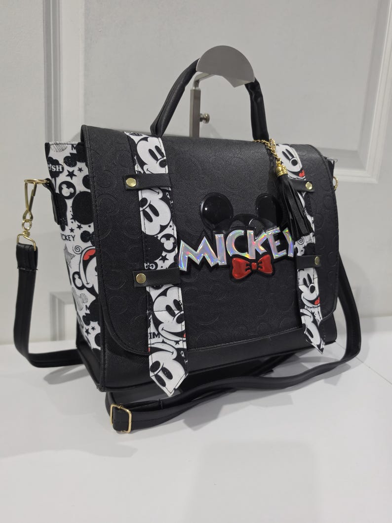 May include: A black satchel bag featuring a debossed pattern, a top handle, and an adjustable shoulder strap. The front flap displays the word MICKEY in iridescent lettering with a red bow, accented by two vertical white strips with black Mickey Mouse graphics.
