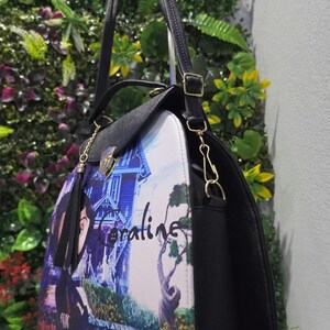 May include: A black handbag featuring a printed graphic of a character and a house with the text Coraline. The bag has a top handle, a detachable shoulder strap with gold-toned metal hardware, a fold-over flap with a clasp, and black side panels.