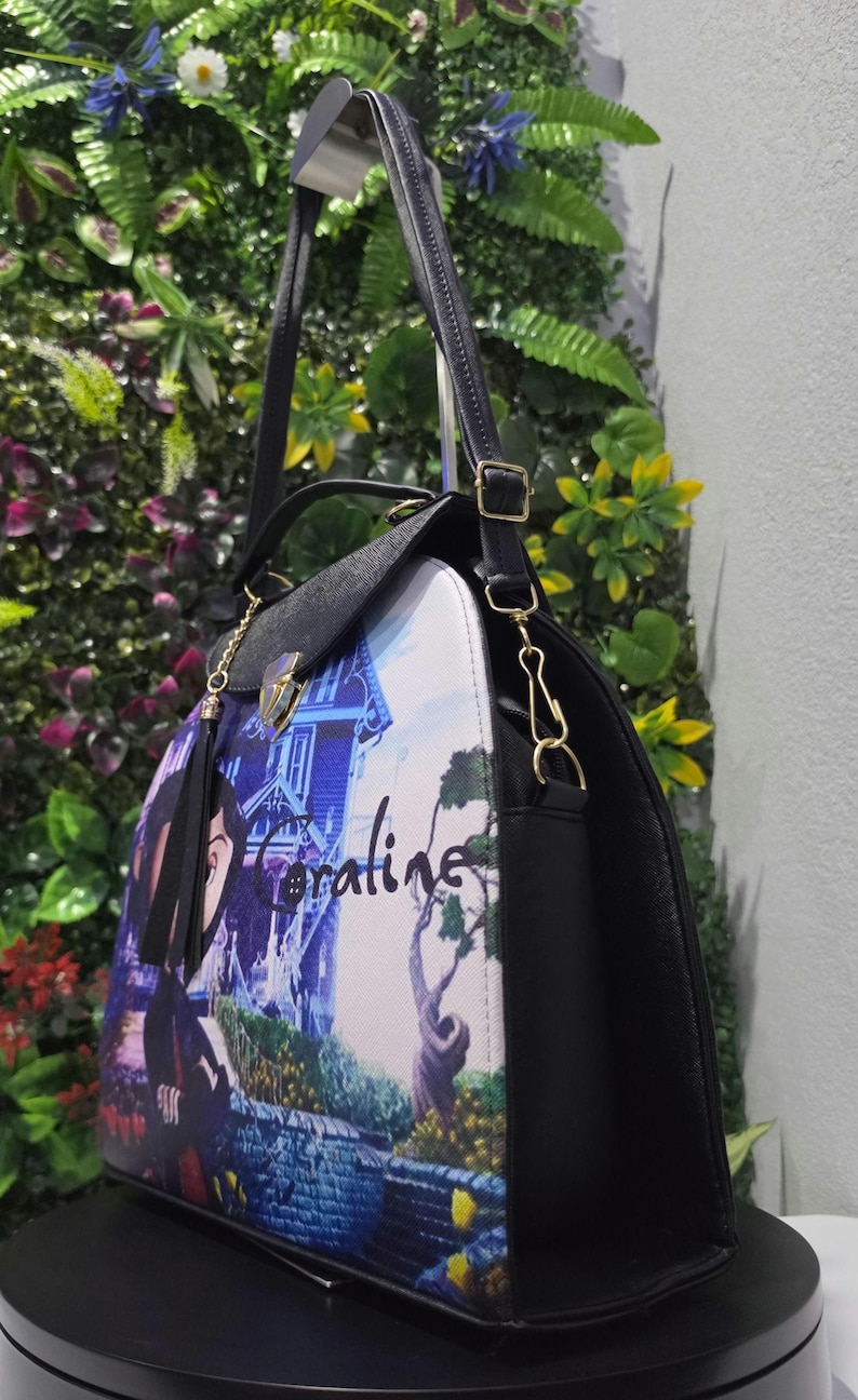 May include: A black handbag featuring a printed graphic of a character and a house with the text Coraline. The bag has a top handle, a detachable shoulder strap with gold-toned metal hardware, a fold-over flap with a clasp, and black side panels.
