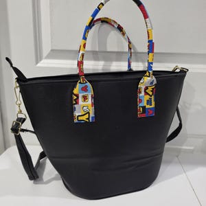 Pu&ograve; includere: Una borsa tote nera con due manici e una tracolla rimovibile. I manici sono rivestiti con un motivo in tessuto colorato con quadrati rossi, gialli e blu, che include sagome di personaggi dei cartoni animati e il testo "M & Y" e "BOY!".