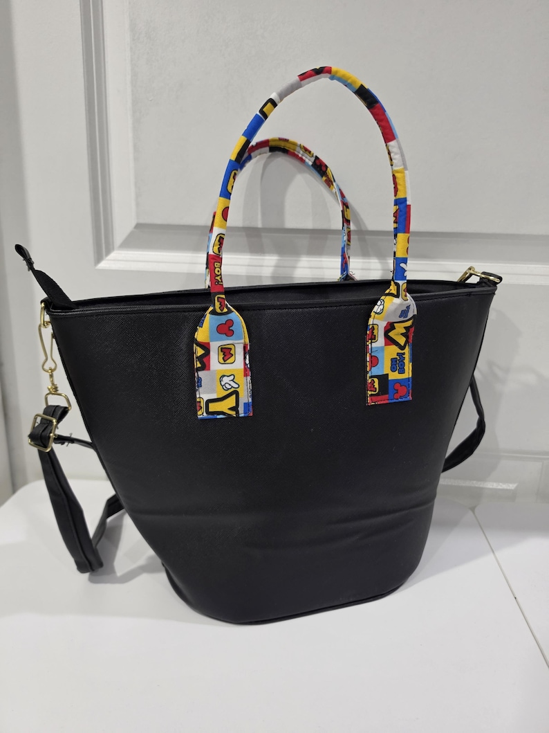 Pu&ograve; includere: Una borsa tote nera con due manici e una tracolla rimovibile. I manici sono rivestiti con un motivo in tessuto colorato con quadrati rossi, gialli e blu, che include sagome di personaggi dei cartoni animati e il testo "M & Y" e "BOY!".