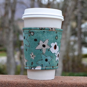 May include: A teal blue and white floral patterned fabric coffee cup cosy with a white paper cup inside.